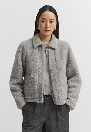 Frau trägt eine hellgraue flauschige Jacke über einem grauen Shirt, mit dunkelgrauen Hosen und goldenen Creolen, steht mit den Händen in den Taschen.