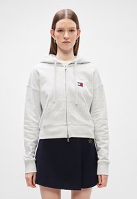 Γκρι φούτερ με φερμουάρ φτιαγμένο από μαλακό ύφασμα, διαθέτει μπροστινή τσέπη, κουκούλα με κορδόνι και ένα μικρό λογότυπο στο στήθος. Συνδυάζεται με μια σκούρα φούστα.