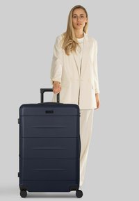 Mujer con traje color crema sostiene el asa de una gran maleta azul marino con ruedas frente a un fondo gris.