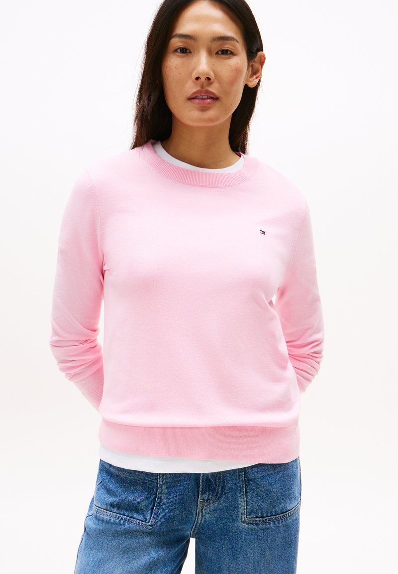 Maglione rosa realizzato in morbido tessuto, con colletto girocollo, maniche lunghe e un piccolo logo sul petto. Indossato con jeans blu.