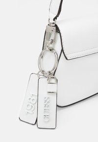 Witte, gestructureerde handtas met zwarte accenten, voorzien van zilverkleurige hardware en twee labels met "1981" en "GUESS." Compacter ontwerp.