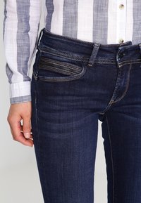 Mörkblå denimjeans med slim fit, featuring en dragkedja, framfickor med texturerade detaljer och kontrasterande sömmar längs sömmarna.