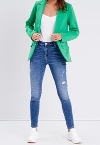 Woman wearing a bright green blazer, white tank top, distressed skinny blue jeans, and white sneakers, standing with hands near blazer sides.