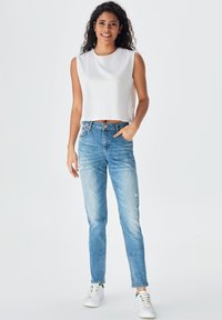 Balta bezpiedurkņu crop top, gaiši zili šaurie džinsi ar nolietojuma detaļām un baltas kedas, izliktas uz vienkārša fona.