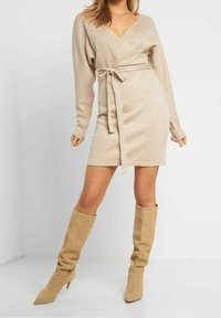 Beige omlottklänning med långa ärmar och djup V-ringning, knuten vid midjan. Bärs med knähöga beige stövlar med spetsig tå.