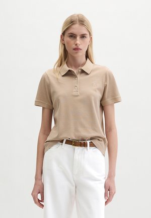 Femme portant un polo beige à manches courtes rentré dans un pantalon blanc taille haute avec une ceinture marron, debout devant un fond uni.