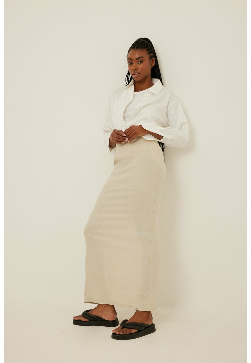 Gonna maxi in lino beige abbinata a una camicia bianca con bottoni, con sandali neri dal design con cinturino, in piedi contro uno sfondo neutro.