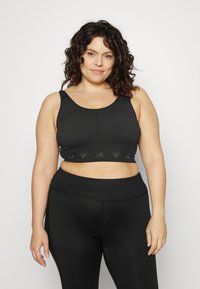 Top desportivo preto com uma superfície texturizada, alças largas e uma faixa elástica com o logótipo da Adidas. Combinado com leggings pretas de cintura alta.