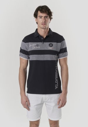 Polo de rayas azul marino y gris con cuello, con texto bordado, logotipo y estampado lateral. Combina con pantalones cortos blancos enrollados.