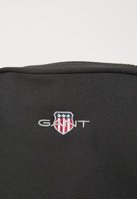 Čierna látková taška s hladkým povrchom, ozdobená výrazným vyšitým logom "GANT" a dizajnom štítu v červenej, bielej a modrej farbe.