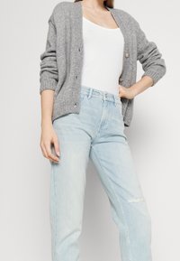 Cardigan gris de punto con puños acanalados y tres botones, usado sobre una camiseta blanca sin mangas y jeans claros rasgados.