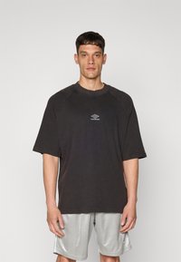 Černé oversized tričko s krátkými rukávy a kulatým výstřihem, na přední straně s bílým logem "Umbro" ve středu. Hladká textilie připomínající bavlnu.