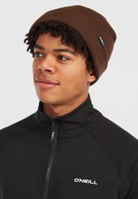 Brun strikket hue med ribbet tekstur, der har et lille grønt logo-tag. Båret sammen med en sort zip-up jakke med hvid branding.