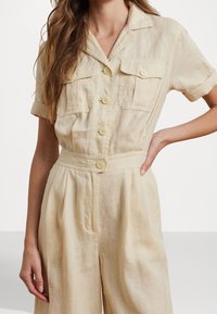 Ljusbeige linnebyxdress med korta ärmar, två bröstfickor, knappstängning fram och veckade vida ben. Mjuk textur.