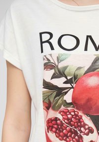Valkoinen puuvillainen lyhythihainen t-paita, jossa on värikäs painatus granaateista ja lehdistä, ja sana "ROME" mustilla lihavoidulla kirjaimilla.