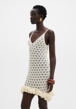 Femeie care poartă o rochie croșetată fără mâneci, de culoare crem, cu tiv cu volane, cercei aurii răsuciți în formă de cerc și două brățări mari maro la încheietura mâinii stângi.