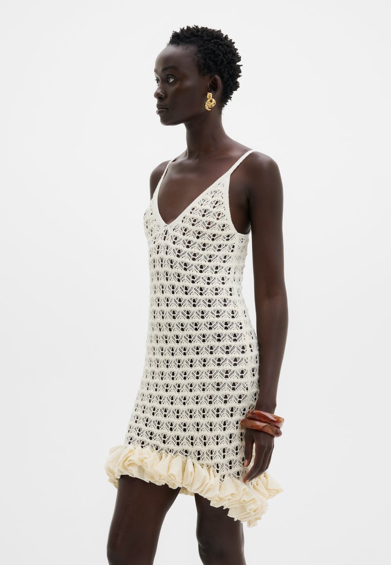 Femme portant une robe sans manches en crochet crème avec un ourlet à volants, des boucles d'oreilles en or torsadé et deux grands bracelets marron au poignet gauche.