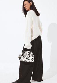 Donna sorridente, che indossa un maglione bianco e pantaloni larghi neri, mentre tiene una borsa bianca con macchie nere e manici neri.