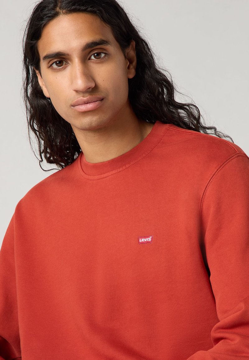 Czerwona bluza z długimi rękawami, okrągłym dekoltem i małą białą naszywką z logo Levi's po lewej stronie klatki piersiowej. Gładka tekstura materiału.