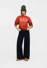 Femme portant un sweat-shirt orange GAP, un pantalon large noir, des chaussures marron et un bonnet tricoté vert, debout les mains dans les poches.