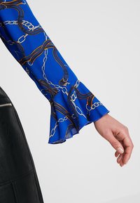 Modrá blůza s rukávem zdobeným vzorem řetězu a pásku, s rozšířeným manžetou. Hladká látka s jemným leskem a kontrastními akcenty.