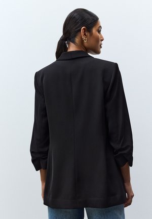 Ženska s temnimi lasmi, ki jih ima spete nazaj, nosi črni blazer s 3/4 rokavi, videna od zadaj na enostavni svetli podlagi.