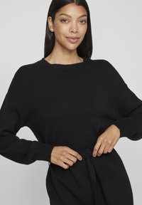 Μαύρος πλεκτός πουλόβερ με χαλαρή εφαρμογή, στρογγυλή λαιμόκοψη και μακριά μανίκια, διαθέτοντας λεπτομέρεια με ζωνάκι στη μέση για ρύθμιση.