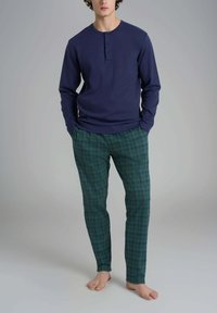 Uomo in piedi a piedi nudi, che indossa una maglietta henley a maniche lunghe blu navy e pantaloni da pigiama a quadri verdi, con le mani nelle tasche.