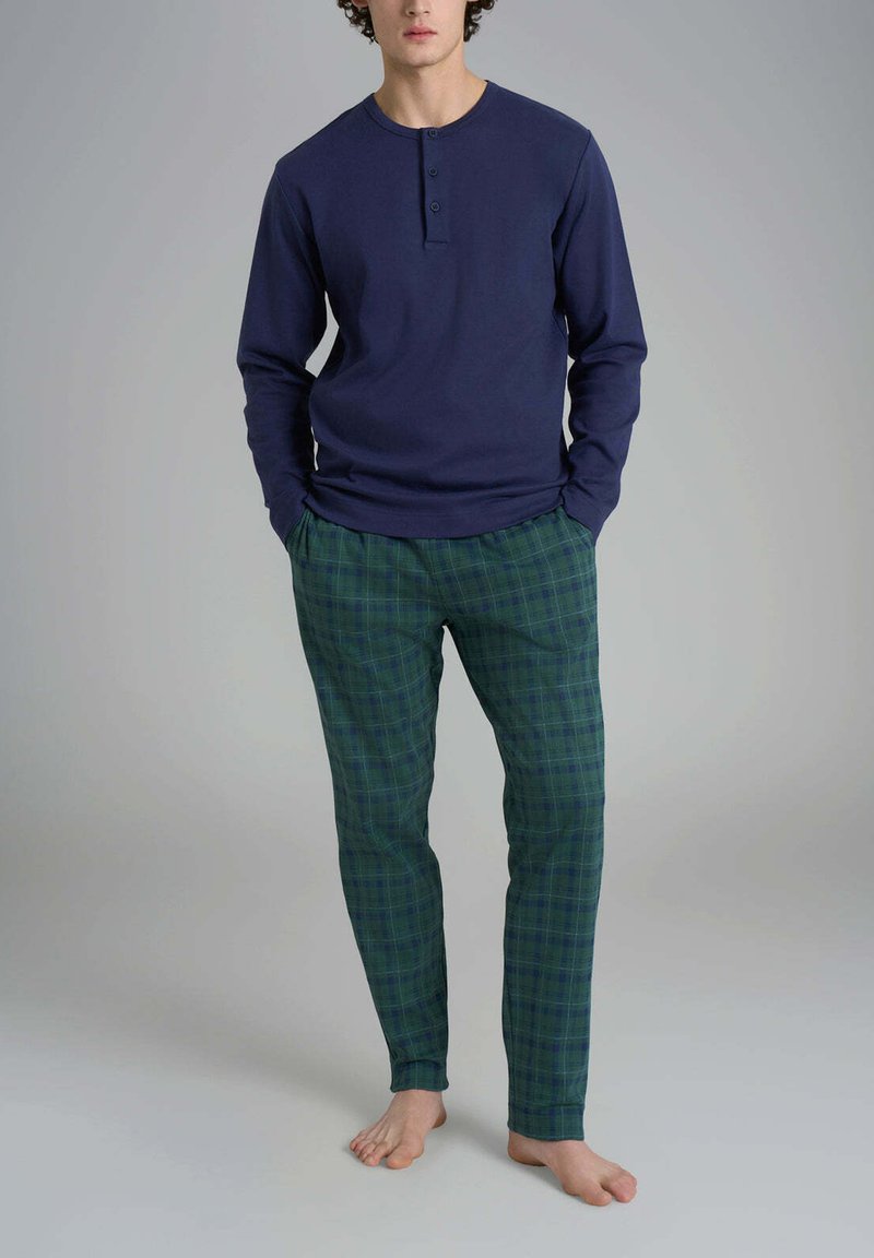 Uomo in piedi a piedi nudi, che indossa una maglietta henley a maniche lunghe blu navy e pantaloni da pigiama a quadri verdi, con le mani nelle tasche.