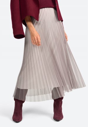 Jupe plissée en tissu taupe doux, avec une finition superposée et transparente. Associée à des bottes au genou bordeaux, mettant en valeur une texture lisse.
