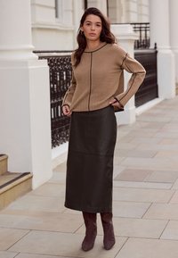 Béžový pletený svetr s černým lemováním, doplněný tmavě zelenou koženou pouzdrovou sukní a hnědými kozačkami nad kolena, stojící venku.