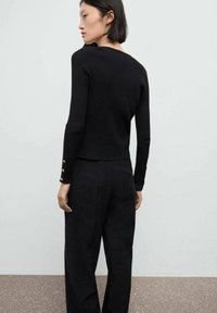 Svart stickad tröja med en figurnära design, ribbad textur och guldknappar som accent på ärmarna. Matchas med svarta byxor, sedd bakifrån.