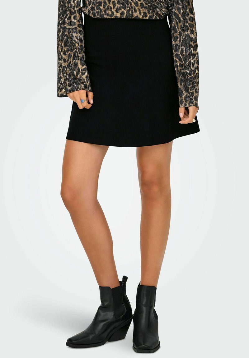 Kobieta w czarnej spódnicy w kształcie litery A, długim rękawie w wzór lamparcze, czarnych botkach za kostkę oraz dwóch pierścionkach na palcach.