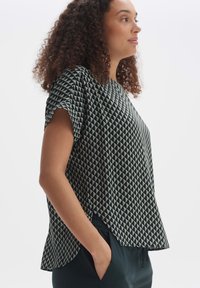 Top a maniche corte di colore verde scuro con un motivo geometrico a triangoli grigio chiaro. Vestibilità rilassata, orlo arrotondato, tessuto morbido.