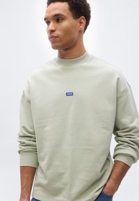 Νέος άνδρας που φοράει ανοιχτοπράσινο φούτερ με μπλε λογότυπο "HUGO", τα χέρια στις τσέπες, κοιτάζοντας αριστερά σε απλό λευκό φόντο.