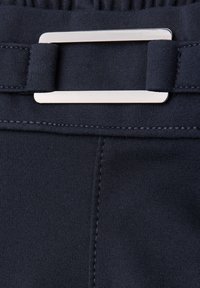 Zbliżenie ciemnej tkaniny z pionowym szwem i elastycznym paskiem w talii, z prostokątną srebrną metalową klamrą.