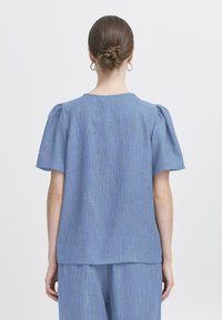 Bluză bleu deschis cu mâneci scurte umflate, având un model texturat cu linii verticale. Guler rotund simplu și croială lejeră.