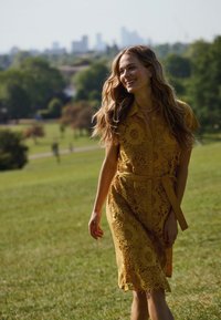 Abito in pizzo giallo senape con motivi floreali, maniche corte, chiusura con bottoni e vita sottolineata da una cintura. Texture morbida, silhouette fluida, ambiente all'aperto.