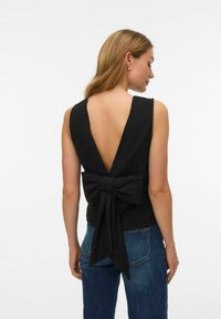 Sieviete valkā bezroku melnu topu ar dziļu V-formas aizmuguri un lielu loku detaļu, kopā ar zilām džinsu biksēm, stāvot priekšā vienkāršam fonam.