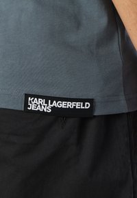 Grå T-shirt med en texturerad yta, försedd med en svart etikett med texten "KARL LAGERFELD JEANS" vid fållen, i kombination med svarta byxor.