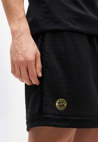 Κοντινό πλάνο ενός χεριού που ακουμπάει δίπλα σε μαύρο αθλητικό σορτς με το λογότυπο της FC Bayern München σε χρυσό χρώμα στο κάτω μέρος.