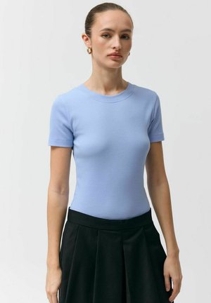 Kobieta ubrana w dopasowany jasnoniebieski krótki rękaw i czarną plisowaną spódnicę z wysokim stanem na jednolitym tle.