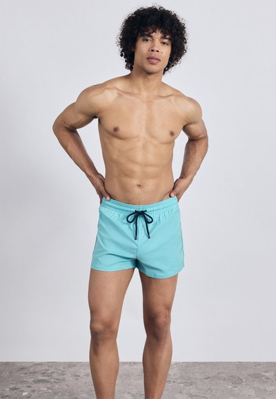 Jauns vīrietis ar cirtaini matiem stāv kaila auguma, ģērbies gaiši zilās peldbiksēs ar melnu auklu uz fona bez dekoriem.