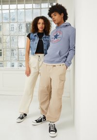 Veste en denim sur un haut noir, pantalon cargo crème, et un sweat à capuche gris avec un logo ; les deux portent des baskets Converse noires ; fond minimaliste.