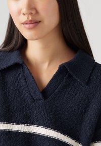 Marinblå stickad tröja med V-ringning och krage, med en vit horisontell rand över bröstet, texturerat tyg.