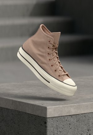 Svetlohnedý vysoký tenisky s bielou gumovou podrážkou a špičkou, vo vzduchu nad textúrovaným sivým betónovým okrajom na rozmazanom pozadí.