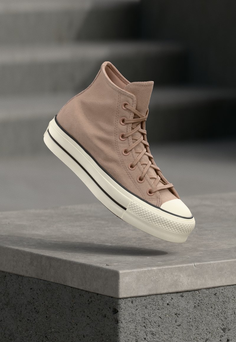 Ανοιχτό καφέ ψηλό αθλητικό παπούτσι με λευκή καουτσούκ σόλα και προστατευτικό στη μύτη, στον αέρα πάνω από μια υφή όψη γκρι σκυροδέματος σε φόντο θολό.