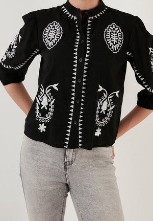 Czarna bluzka na guziki z białym kwiatowym i geometrycznym haftem na piersi, rękawach i przednich krawędziach, w zestawieniu z jasnoszarymi dżinsami.