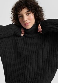 Zwarte coltrui met een grove gebreide textuur en een losse pasvorm. Voorzien van dikke geribbelde manchetten en een hoge hals voor extra warmte.