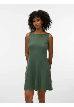 Kobieta o kręconych włosach, ubrana w zieloną, bezrękawnikową sukienkę sięgającą kolan, stoi z rękami za plecami na jednolitym białym tle.